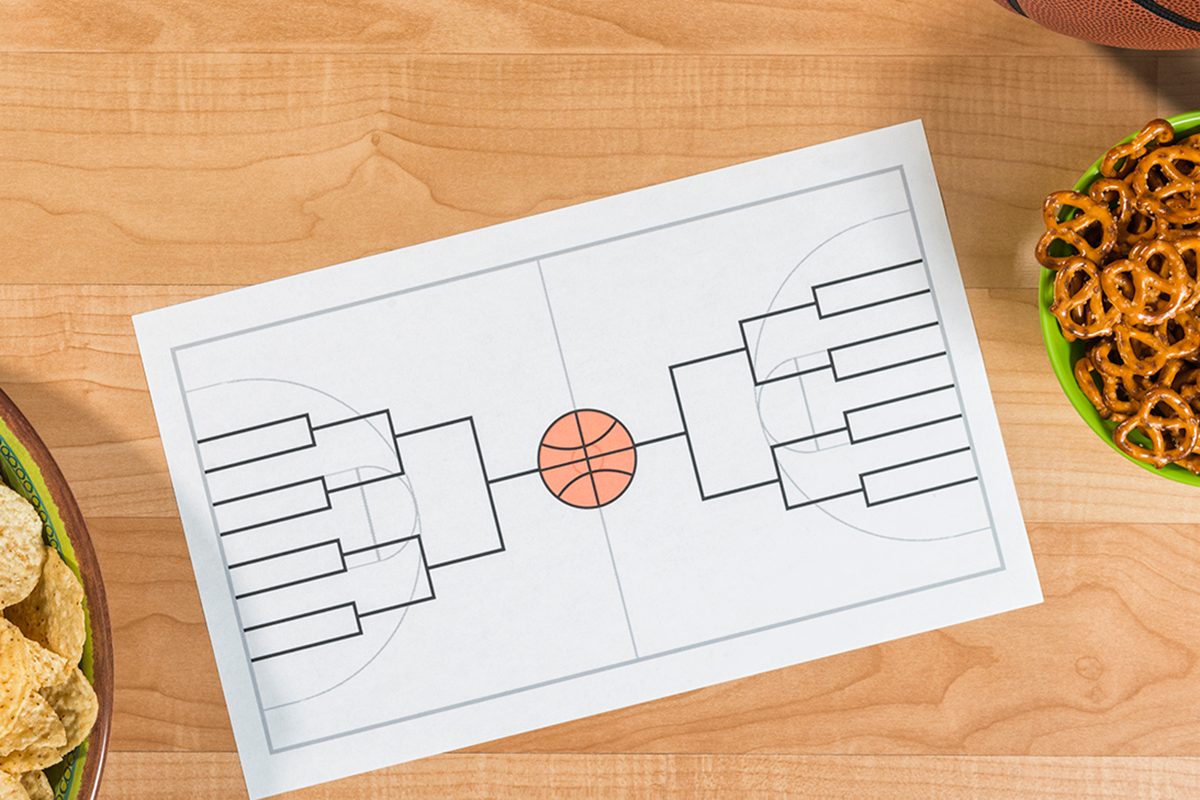 An overhead view of a party for a college basketball tournament with an empty 16 team bracket. There is a bowl of small pretzels, chips and salsa along with a basketball sitting on what looks like a maple wood basketball court. This bracket is for a 16 team single elimination tournament, winner take all, which typically happens in March every year in the US between college basketball teams and many people gamble on the series.