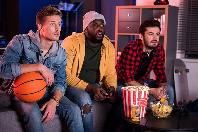 So exciting. Three young interested guys are sitting on sofa and expressing exhilaration while watching sport game with opened mouthes. Friends are eating popcorn and drinking beer