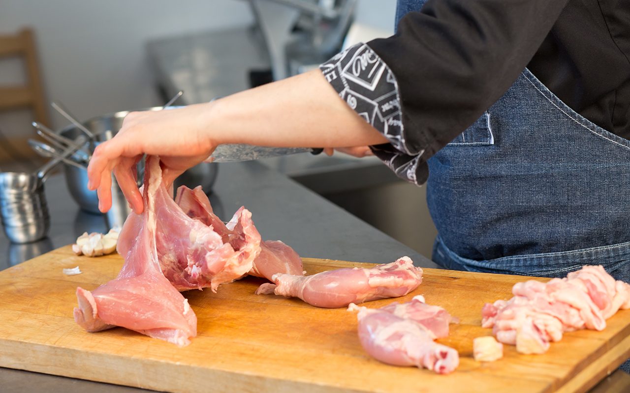 A person cuts raw chicken. Cook's hand with a knife close-up on the background of the kitchen. the woman professional chef holds raw chicken. The background is blurred