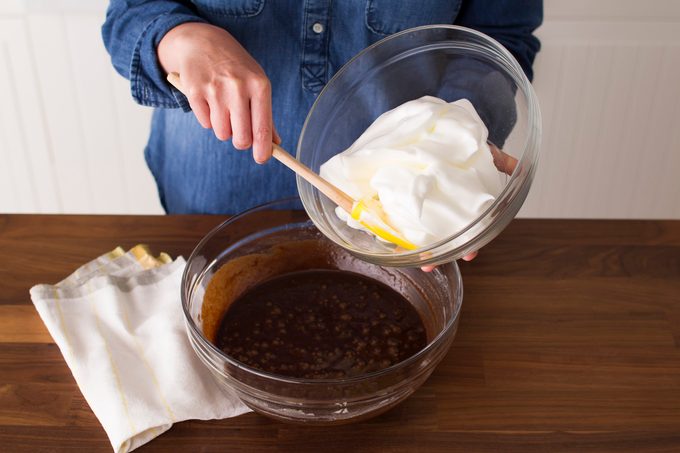 Person digging into a bowl of whipped cream with a spatula while standing over a bowl of melted chocolate