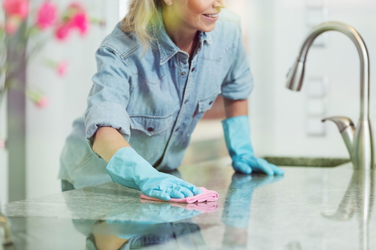 Woman wiping down kitchen countertop with pink cloth, wearing blue jean shirt and rubber gloves for household cleaning