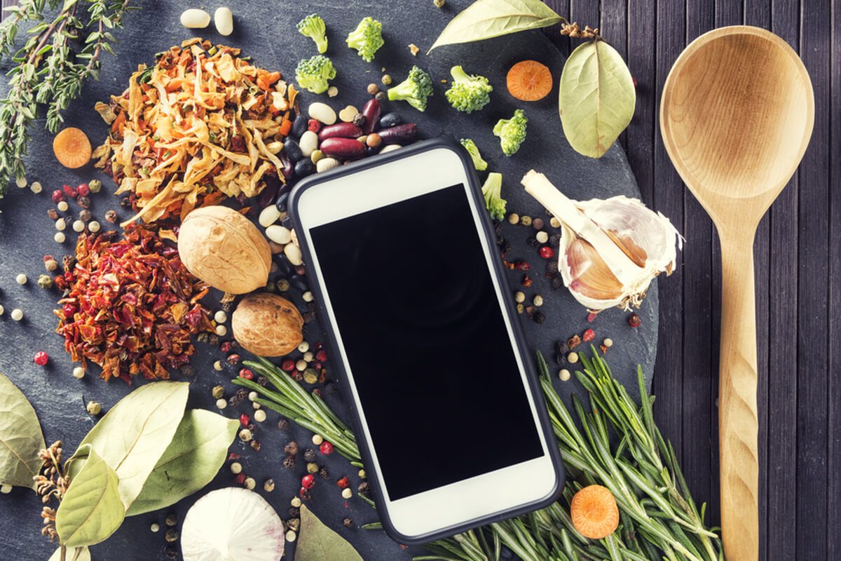 Kitchen table with ingredients, utensils and smartphone with blank screen for your app over cooking book on wooden table.