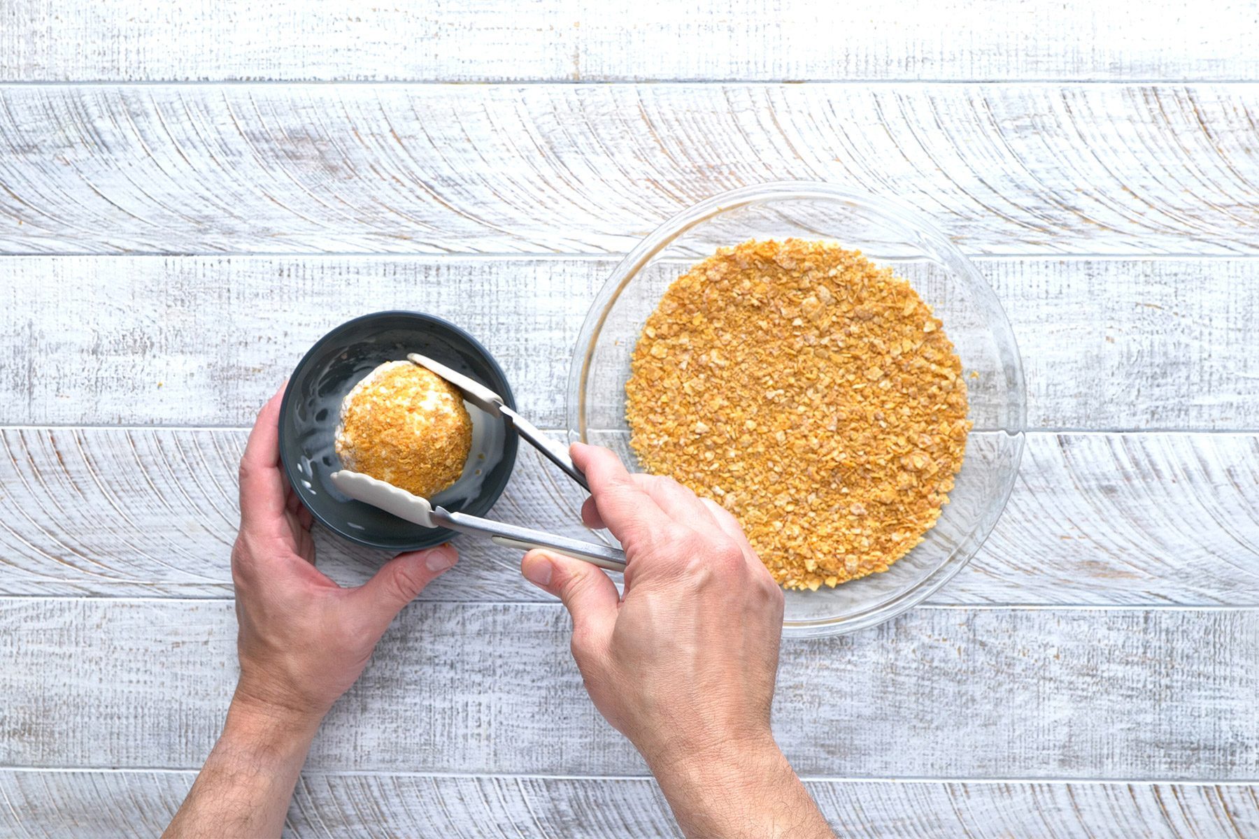 overhead shot; white wooden background; placing rolled ice cream scoop in cornflake mixture in a small bowl; cornflake mixture in a plate