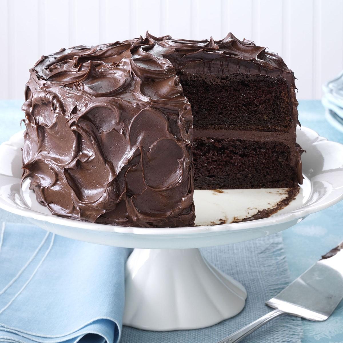 A chocolate layer cake with rich, swirled chocolate frosting sits on a white cake stand. A slice is cut, revealing the moist, dark interior. The cake is set on a blue cloth with a knife nearby.