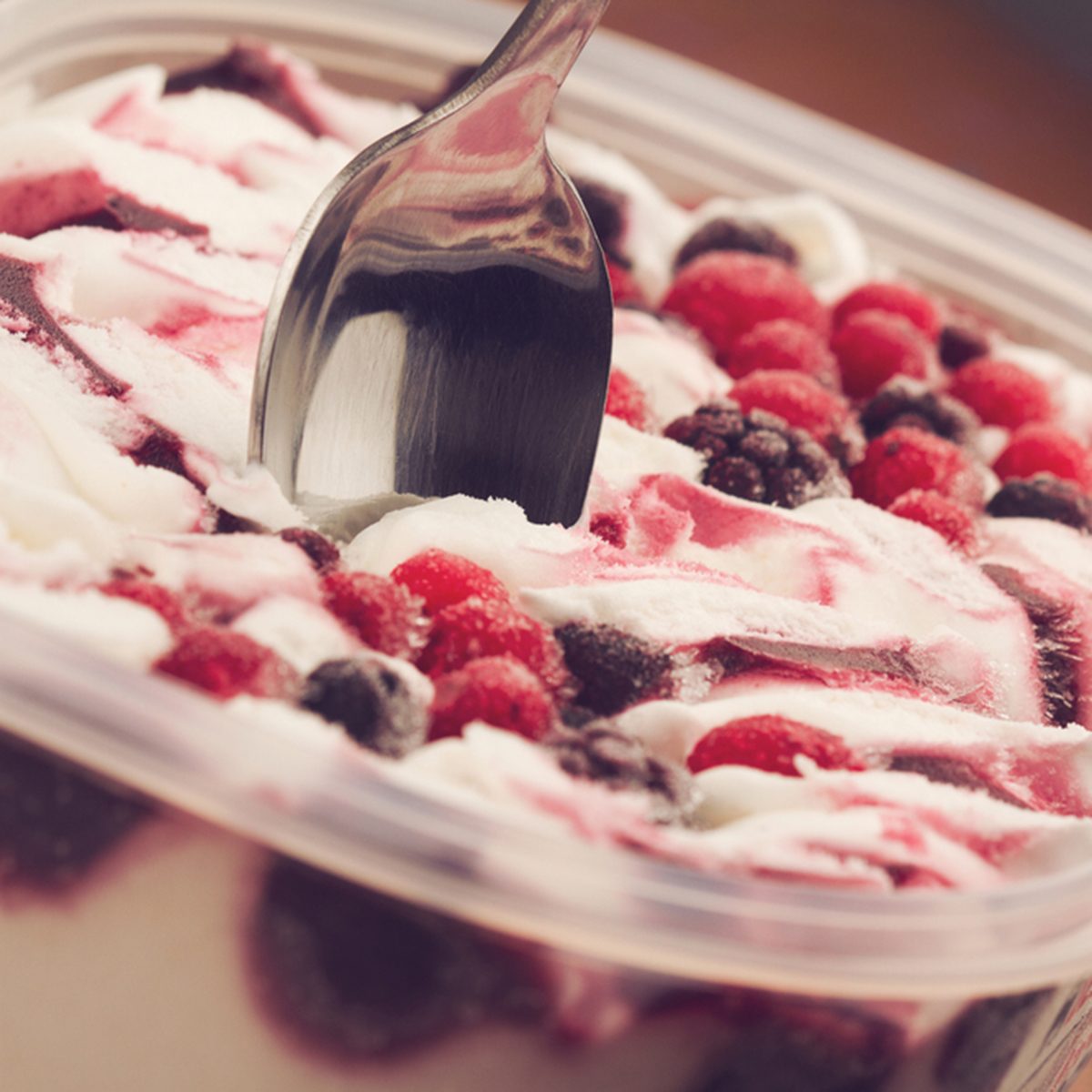 horizontal close up of chrome teaspoon stuck in vanilla ice cream with mix of black and red frozen berries on brown background selective focus