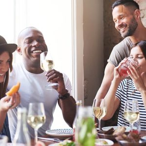 Gathering of four diverse laughing adults enjoying wine and appetizers at lunch with each other at restaurant table beside large bright window