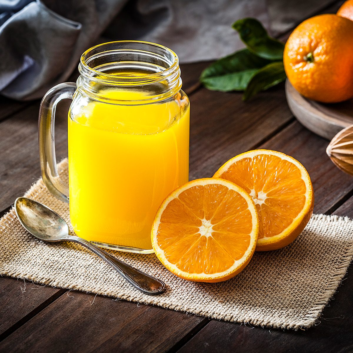 Orange juice glass jar shot on rustic wooden table. The jar is on a burlap cloth and two orange halves are beside it. An old metal spoon and a wooden juicer complete the composition. A round wooden tray with fresh oranges is at the top-right corner of an horizontal frame. Predominant colors are orange and brown. DSRL studio photo taken with Canon EOS 5D Mk II and Canon EF 100mm f/2.8L Macro IS USM
