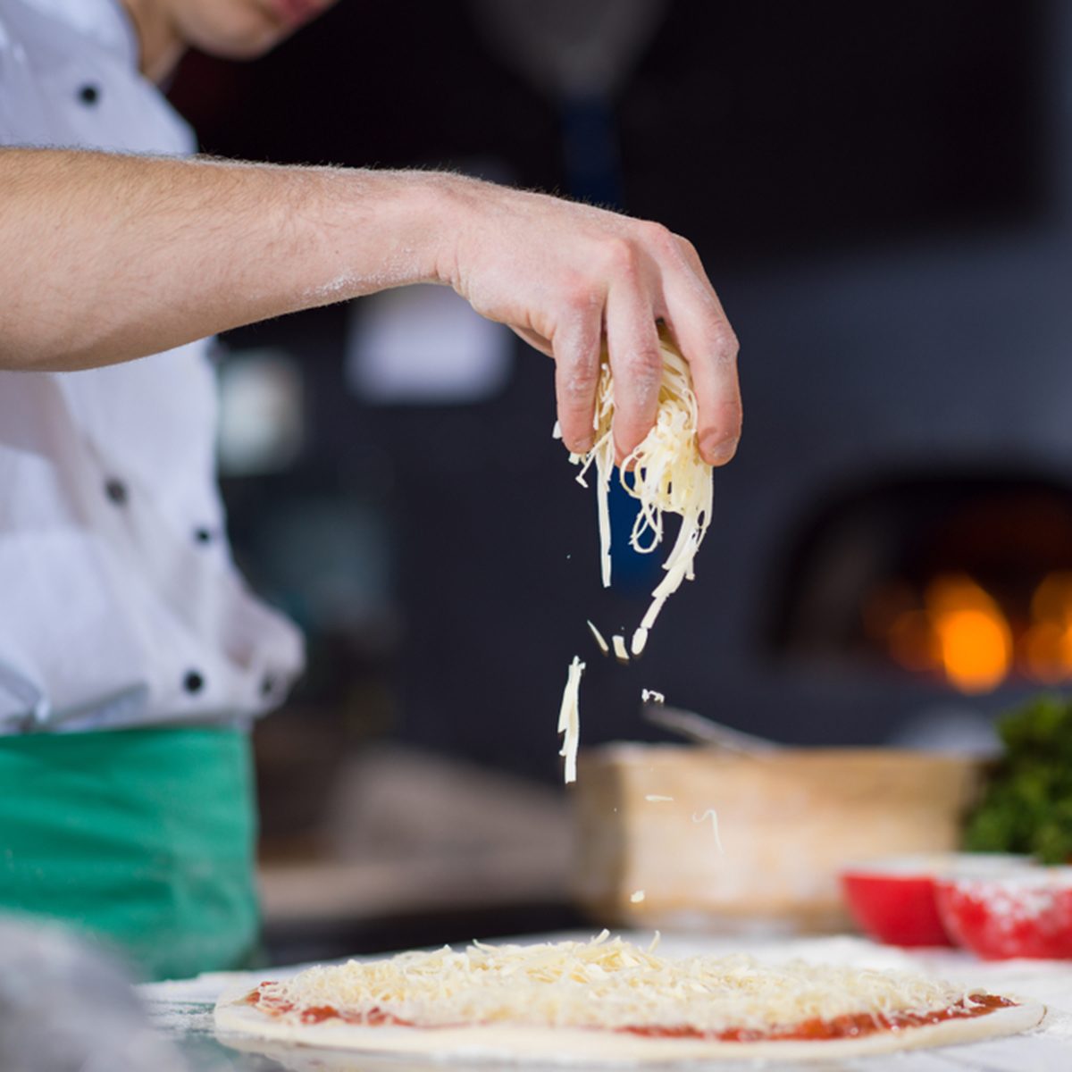 chef sprinkling cheese over fresh pizza dough on kitchen table