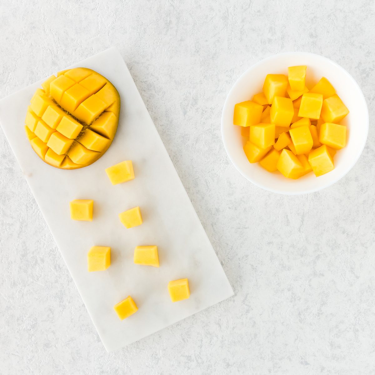 Top view of cut mango half and mango cubes on white marble cutting board and in white bowl.