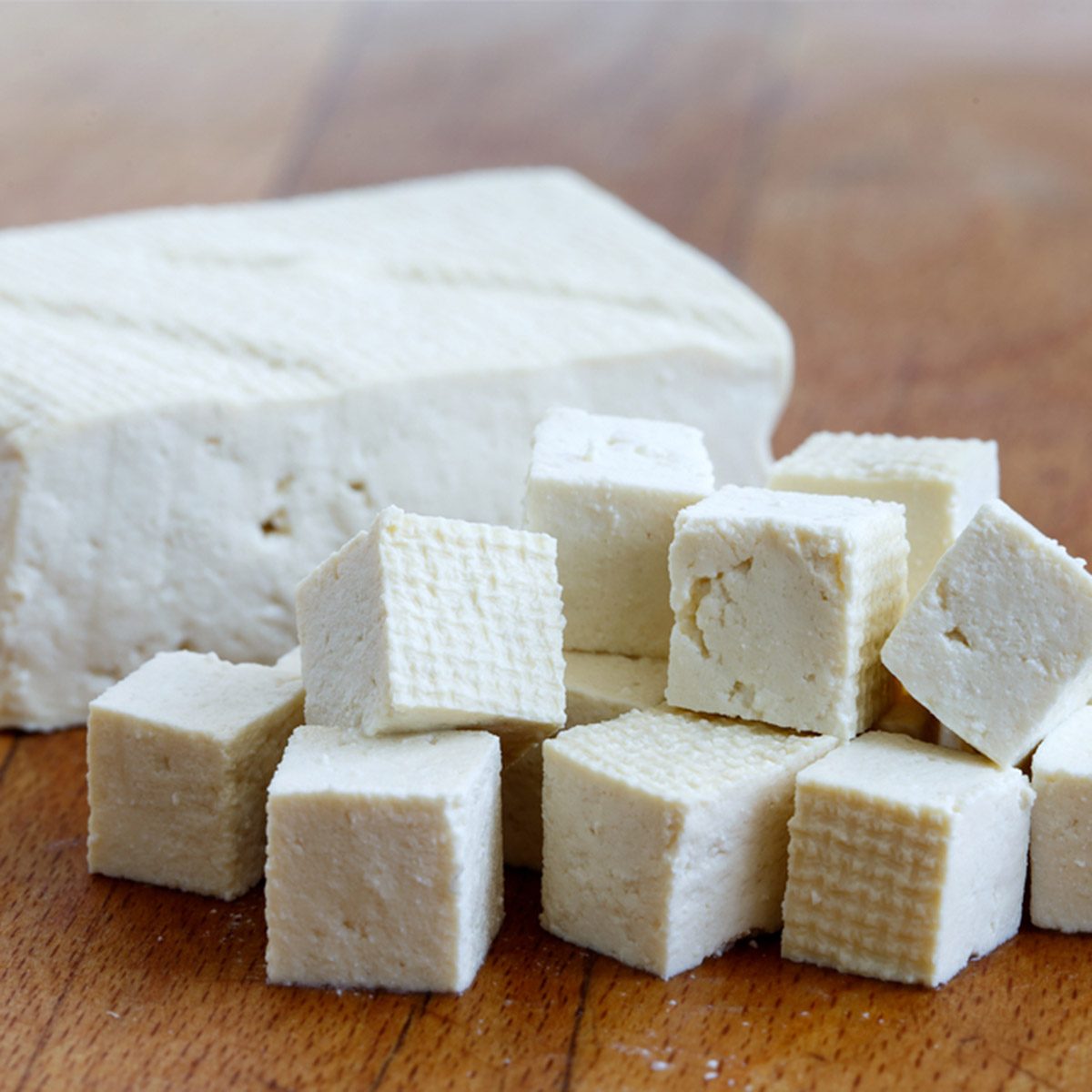 Single block of white tofu with cut tofu cubes on wooden chopping board.