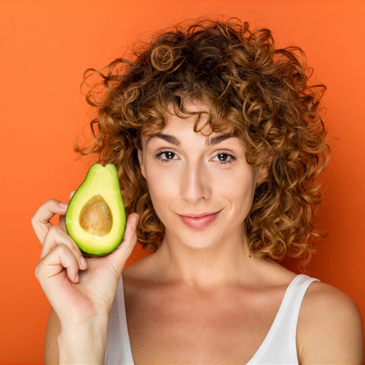 young curly woman holding a avocado in hands on orange background