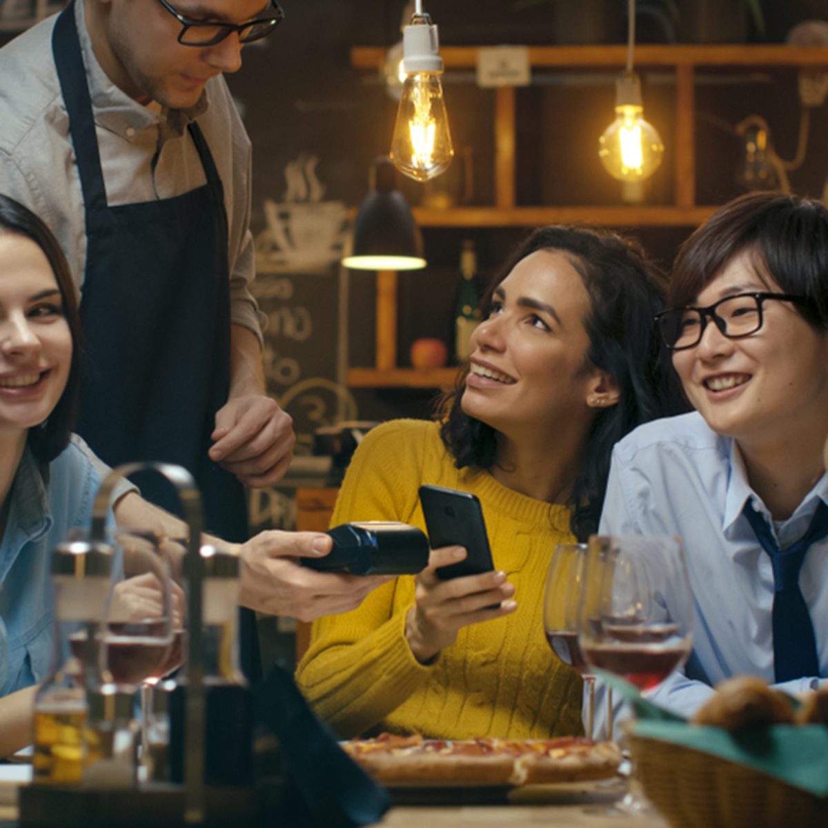 In the Bar Waiter Holds Credit Card Payment Machine and Beautiful Woman Pays for Her Order with Contactless Mobile Phone Payments System.