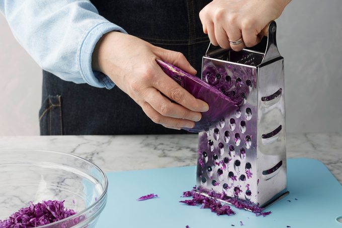Cabbage Being Grated with a box grater over Blue Cutting Board