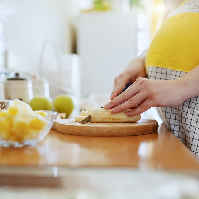 Close up of Caucasian pregnant woman in apron cutting banana and preparing healthy breakfast.