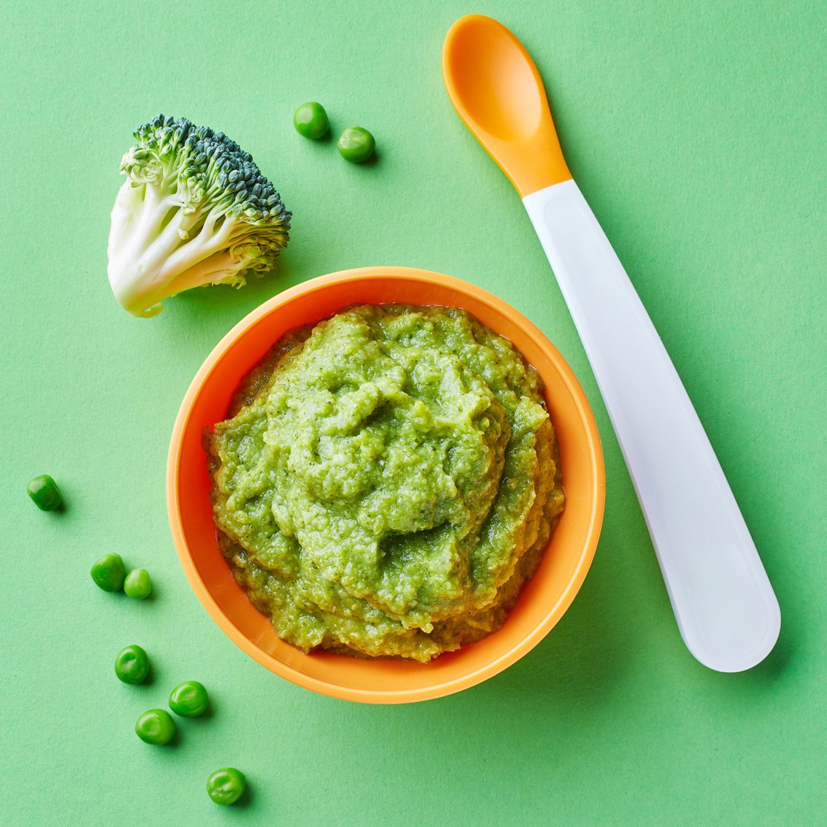 Green peas and broccoli baby puree in bowl with baby spoon on green background