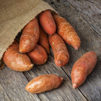 Freshly harvested organic sweet potatoes spilling from a burlap bag onto a natural weathered wood table.