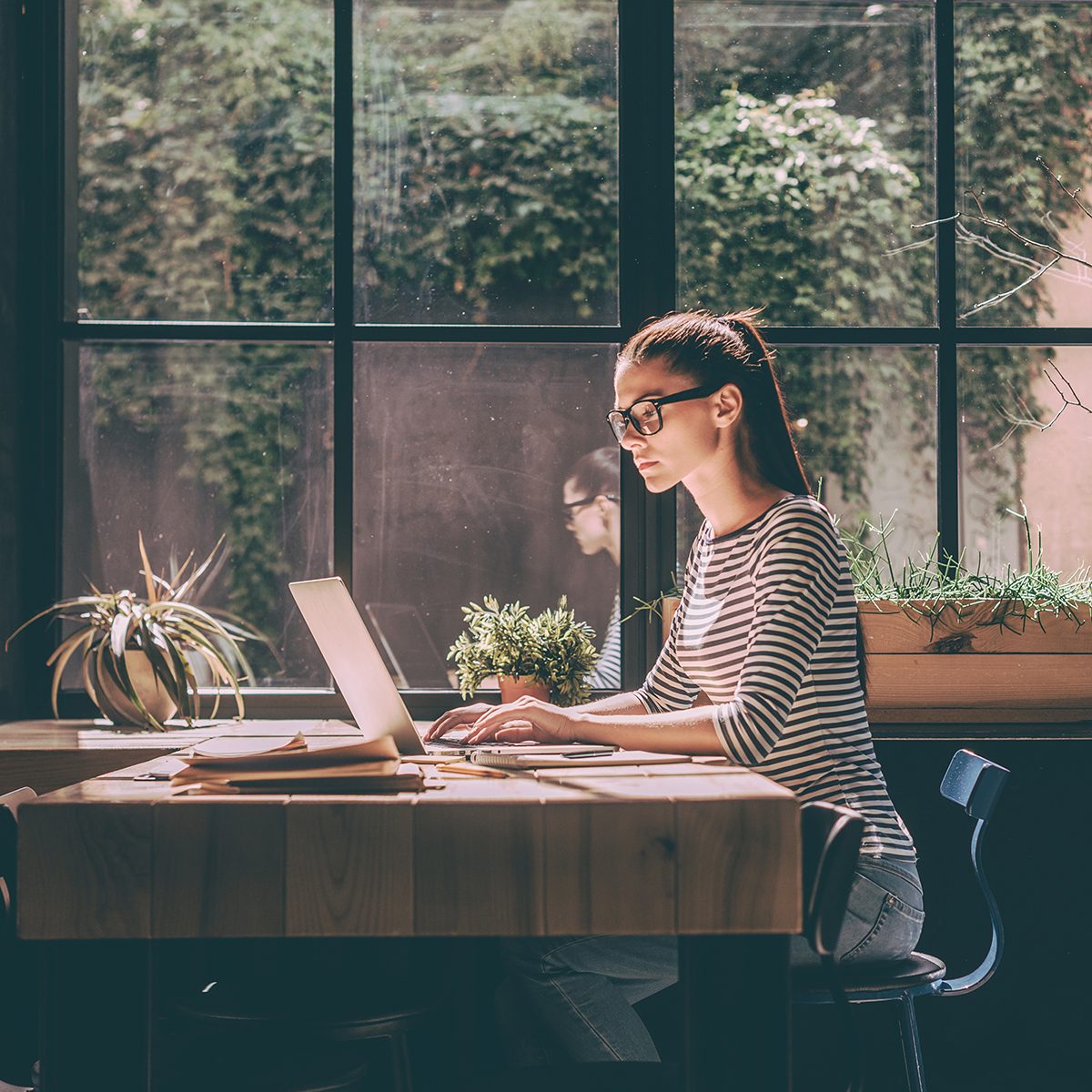 Concentrated at work. Confident young woman in smart casual wear working on laptop while sitting near window in creative office or cafe