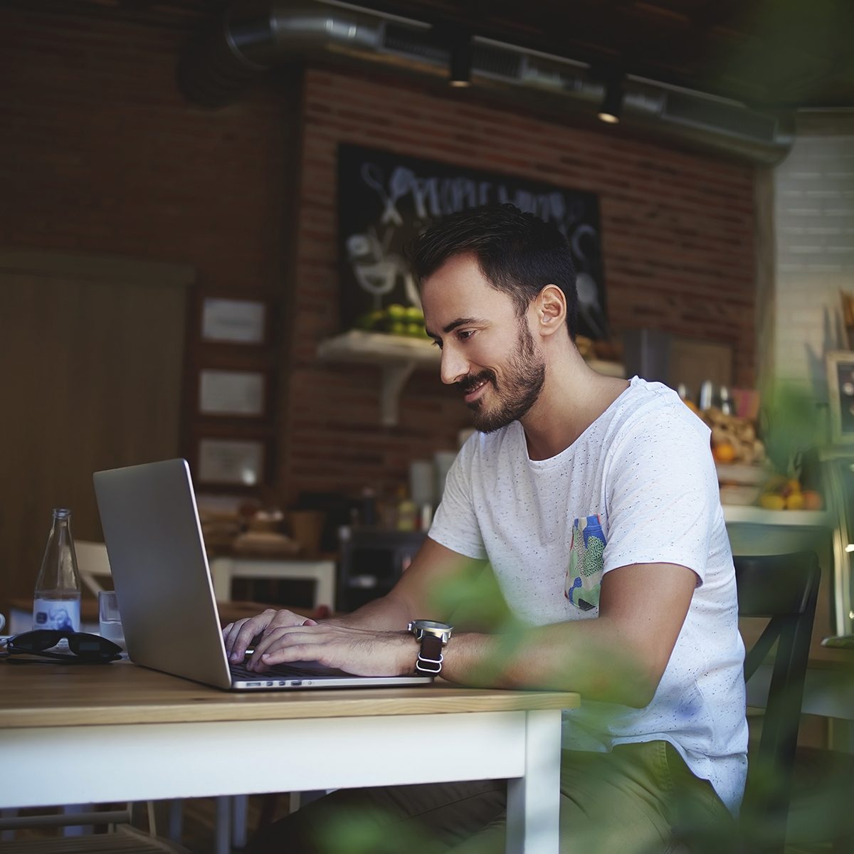 Smiling Caucasian man rewriting in social network with his friend via laptop computer while resting in bakery store, happy male student writing text on net-book during breakfast in modern coffee shop