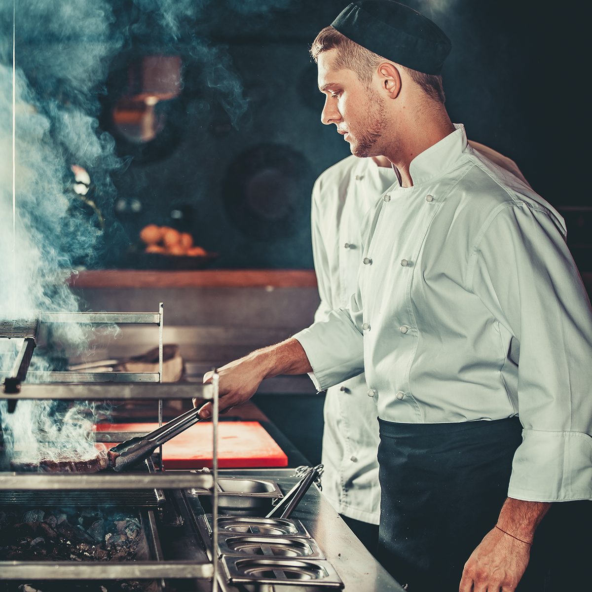 Young white chef in black apron and hat standing near the brazier whith coals. 