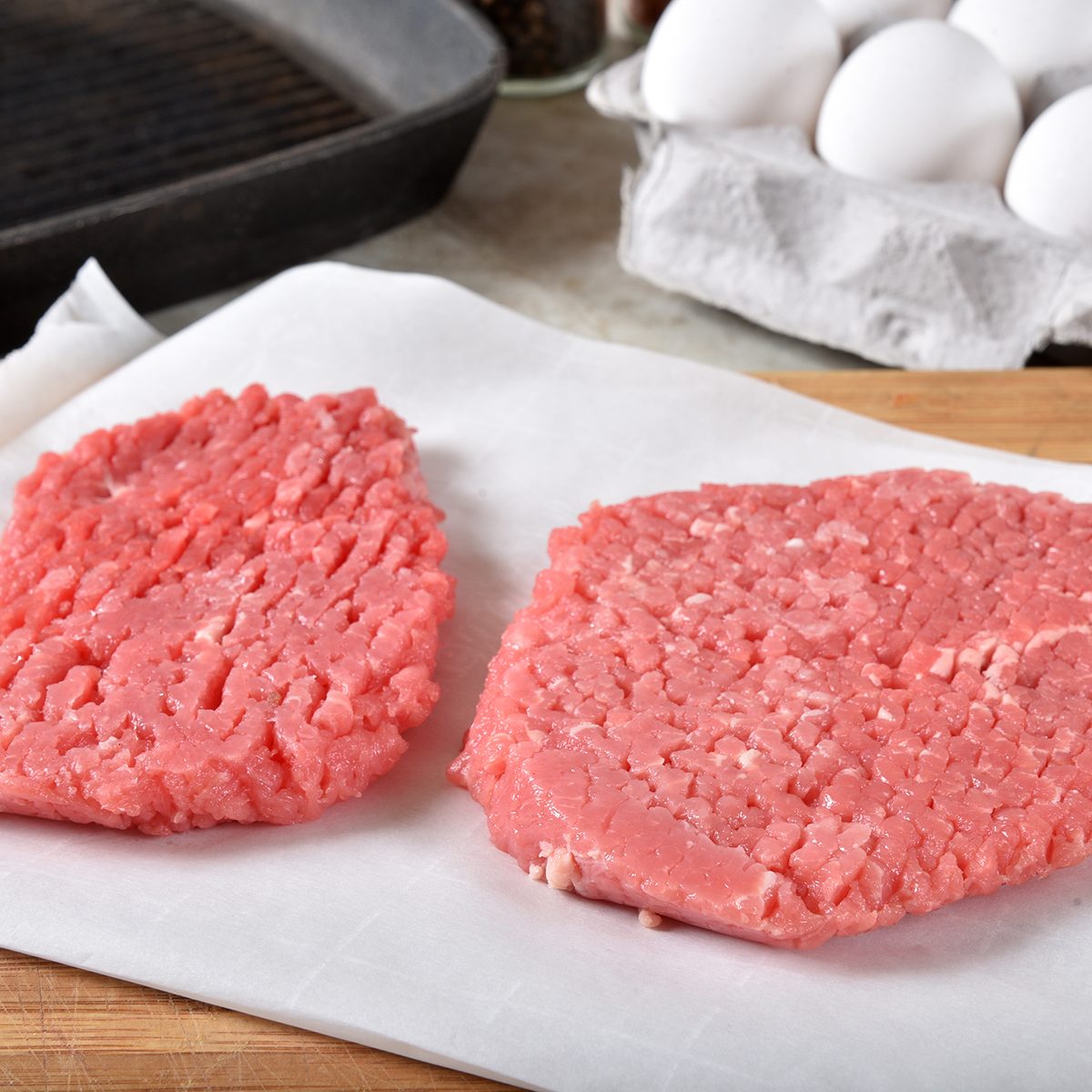 Uncooked cube steaks on a cutting board near a cast iron grill and a carton of eggs
