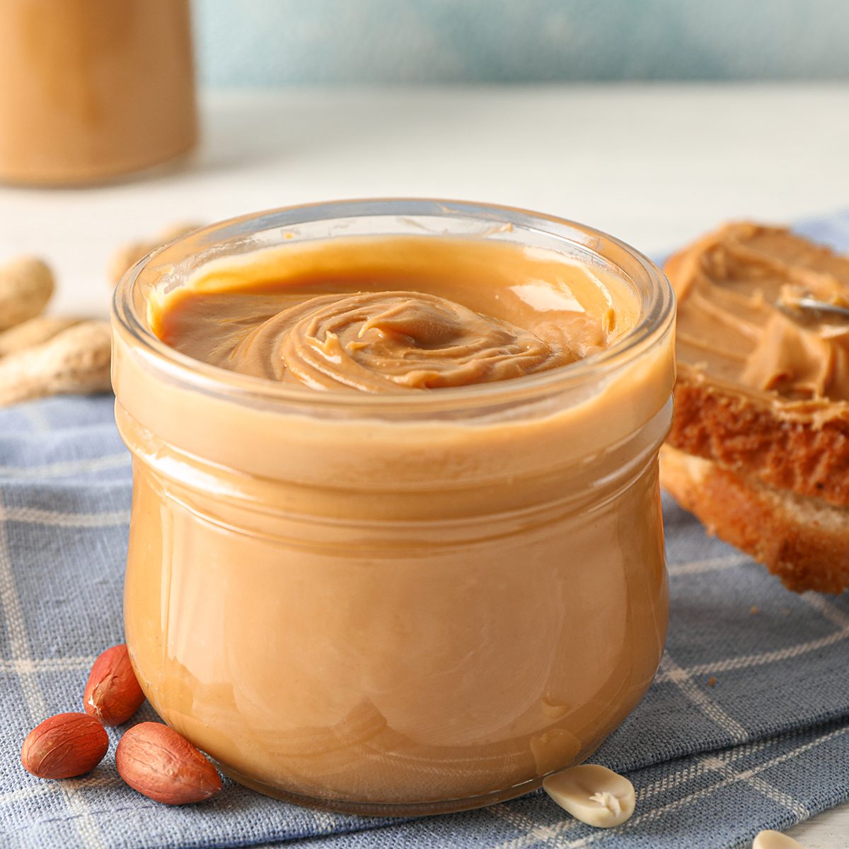 Glass jar with peanut butter, peanut, kitchen towel, spoon and peanut butter sandwich on white wooden background