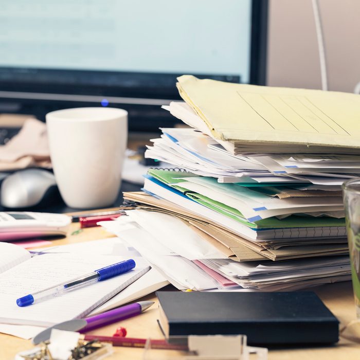 stack of brochures, files and documents, on mess desktop, monitor of pc in background