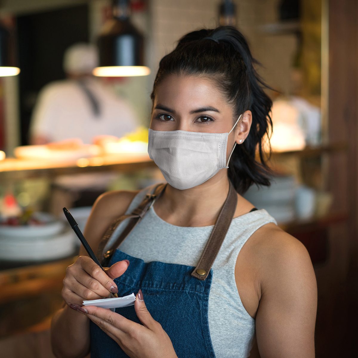 Portrait of a beautiful waitress working at a restaurant wearing a facemask while holding a notepad and looking at the camera – pandemic lifestyle concepts