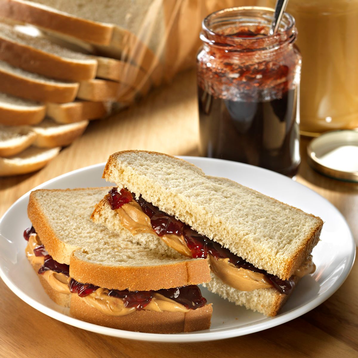 Peanut Butter and Jam sandwich on whole grain white bread with peanut butter jar and jam jar in background with loaf of bread and bottle of milk.