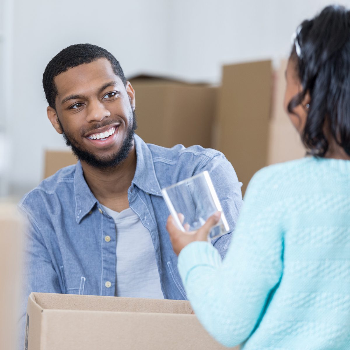 Little girl holds a drinking glass while unpacking a box in her new home. Her back is to the camera. Her dad is smiling at her.