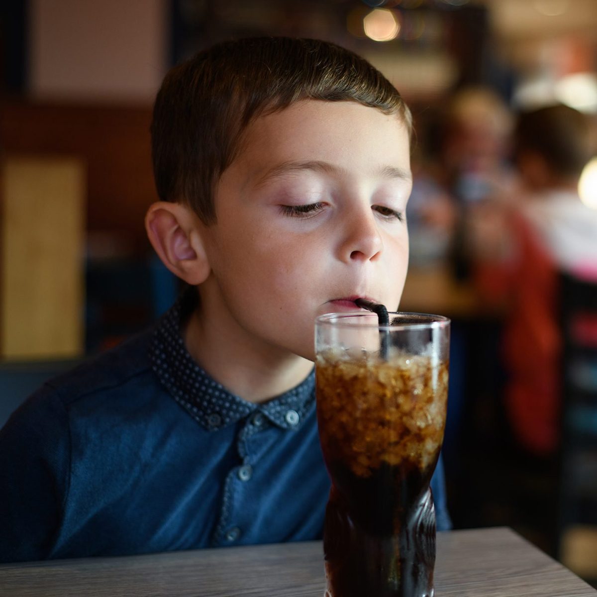 Image of a six year old boy drinking a sugary drink (cola) through a straw sitting at a table in a restaurant.