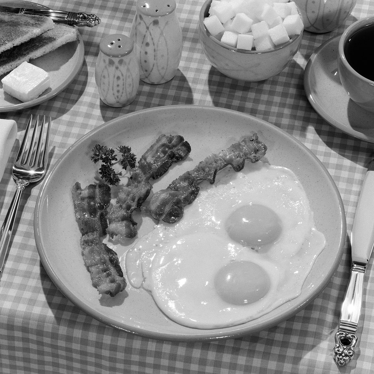 1950s BREAKFAST PLATE OF BACON AND FRIED EGGS AND COFFEE SUGAR BOWL TOAST SALT AND PEPPER SHAKERS ON CHECKERED TABLECLOTH (Photo by L. Fritz/ClassicStock/Getty Images)
