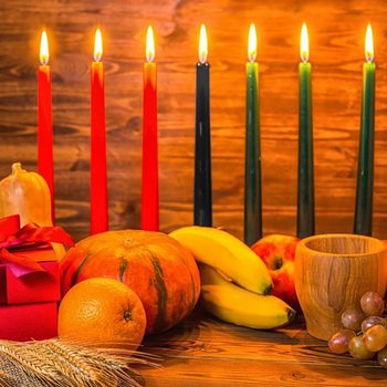 Kwanzaa holiday concept with traditional lit candles, gift box, pumpkins, ears of wheat, grapes, orange, banana, bowl and fruits on wood background