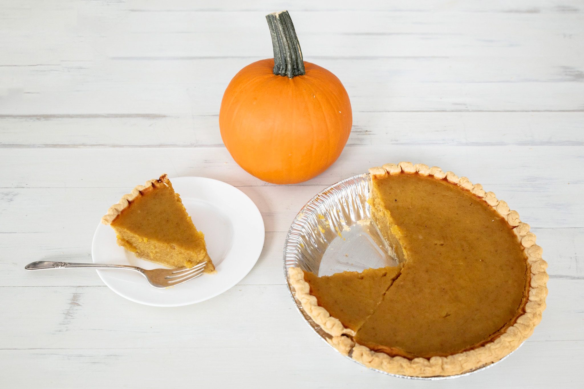 a slice of pumpkin pie on a plate with a fork, a small pumpkin and a pie dish with a pumpkin pie with a missing slice