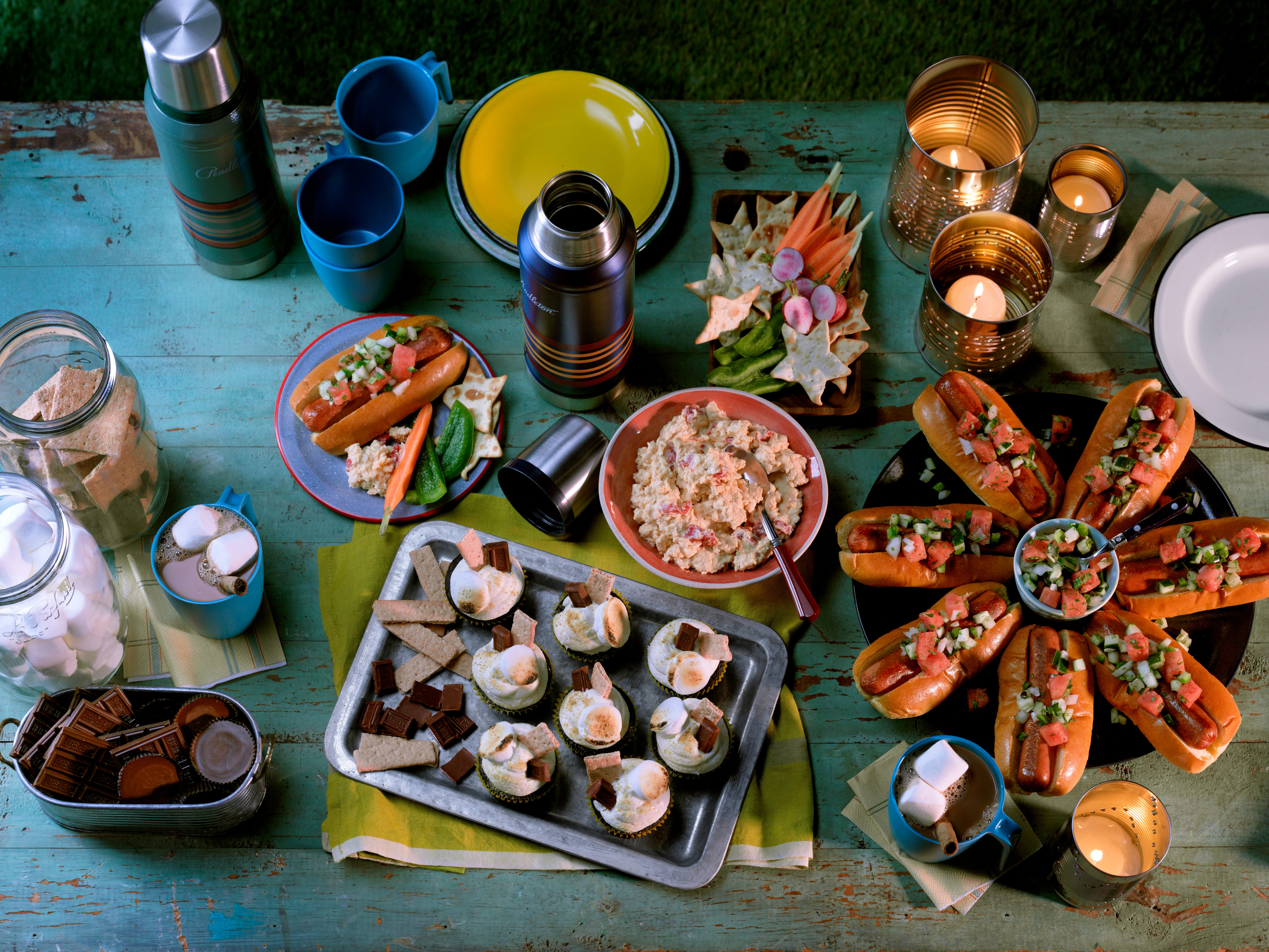 PU5534; opener; HCA opener; Dusk; dusky; night; yard; backyard; summer; picnic; nioght; party; bonfire; overhead camera angle; wood surface blue wood surface; green wood surface; picnic table; outdoors; 16461 Favorite Hot Chocolate; 42362 S'mores Cupcakes; 112361 Cheese and Pimiento Spread; 167568 Watermelon and Cucumber Salsa; hotdogs; buns; candles; thermos; s'mores; marshmallows; chocolate; graham crackers; cocoa; veggie tray; veggies; wood tray; copy space; 2 page spread; condensed milk; baking cocoa; water; vanilla extract; Sweetened whipped cream; marshmallows; chocolate syrup; Pirouette cookies; sharp white cheddar cheese; cream cheese; Worcestershire sauce; white vinegar; white pepper; garlic powder; cayenne pepper; pimientos; crackers; vegetables; veggieswater; buttermilk; eggs; canola oil; vanilla extract; flour; sugar; baking cocoa; baking soda; salt; baking powder; FROSTING; butter; confectioners' sugar; vanilla extract; marshmallow creme; graham cracker crumbs; milk chocolate candy bars; Toasted marshmallows; watermelon; chopped cucumber; sweet onion; fresh cilantro; jalapeno pepper; lime juice; salt; salsa
