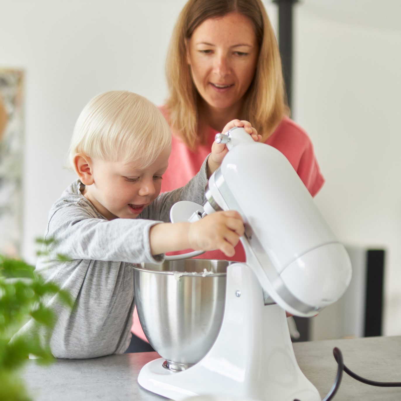 A mother bakes bread with her little blond-haired son in her modern kitchen and teaches him how to operate the food processor and wash his hands properly after having finished the dough.