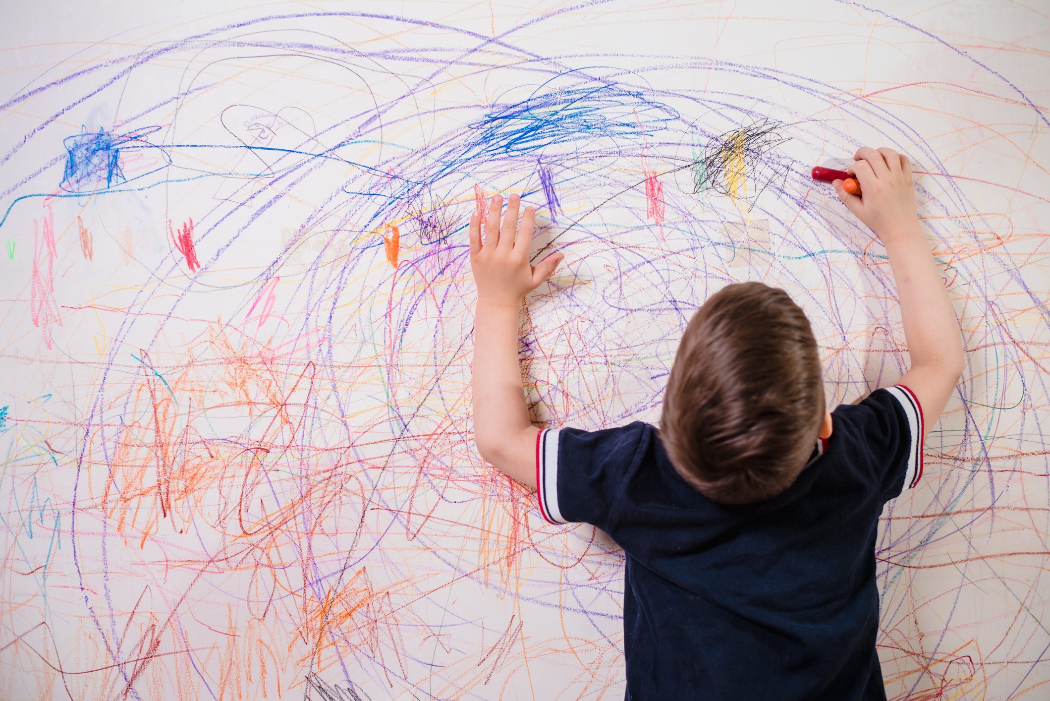 The Child Draws On The Wall With A Crayon. The Boy Is Engaged In Creativity At Home