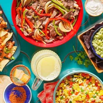 overhead view of colorful tex-mex food, nachos, fajitas, chips and guacamole, corn salsa, and a pitcher of margaritas on a teal tiled table