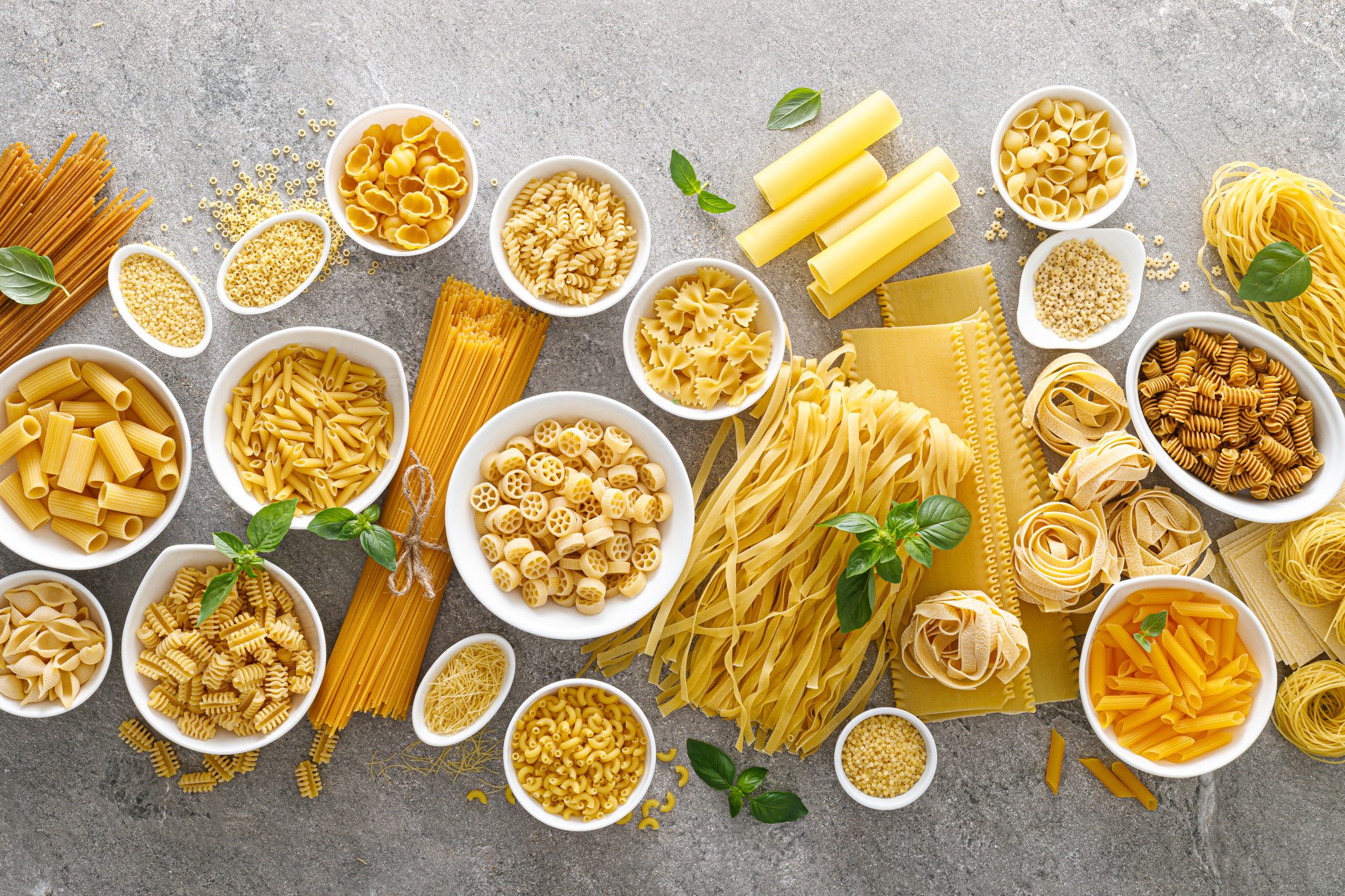 Various kinds of uncooked pasta and noodles, some in white bowls, on stone background, top view