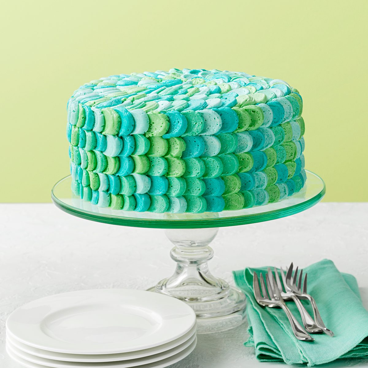 A cake with a decorative icing pattern in shades of blue and green sits on a glass cake stand. The background is light green, and nearby are stacked white plates and a folded green napkin with forks on top.