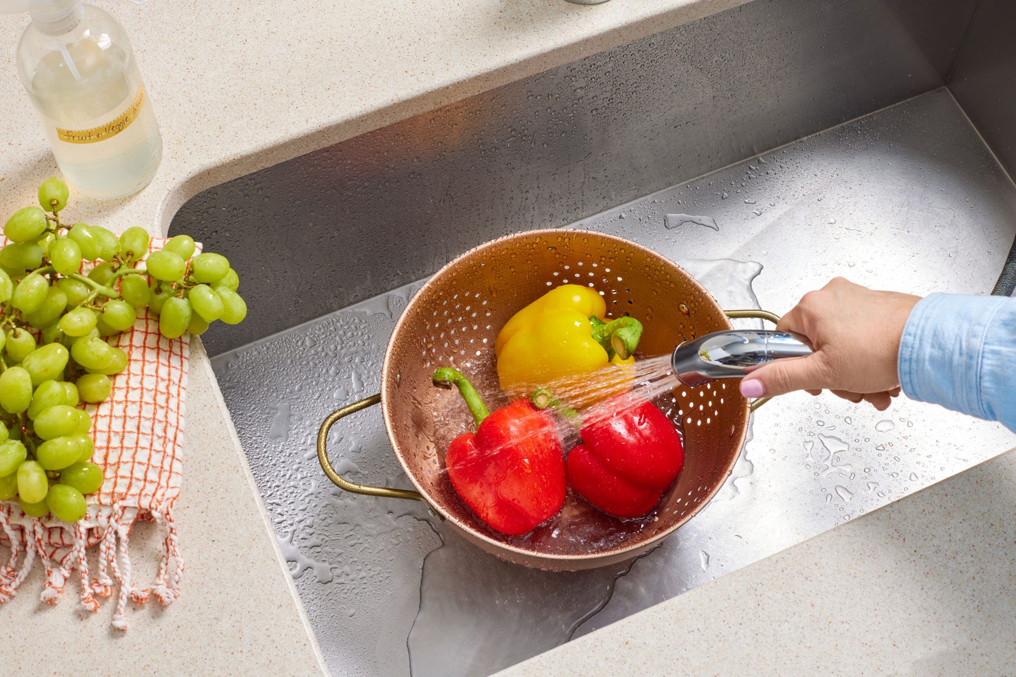 three bell peppers are in a colander in a stainless steel sink. a hand uses the sprayer faucet to rinse the them..