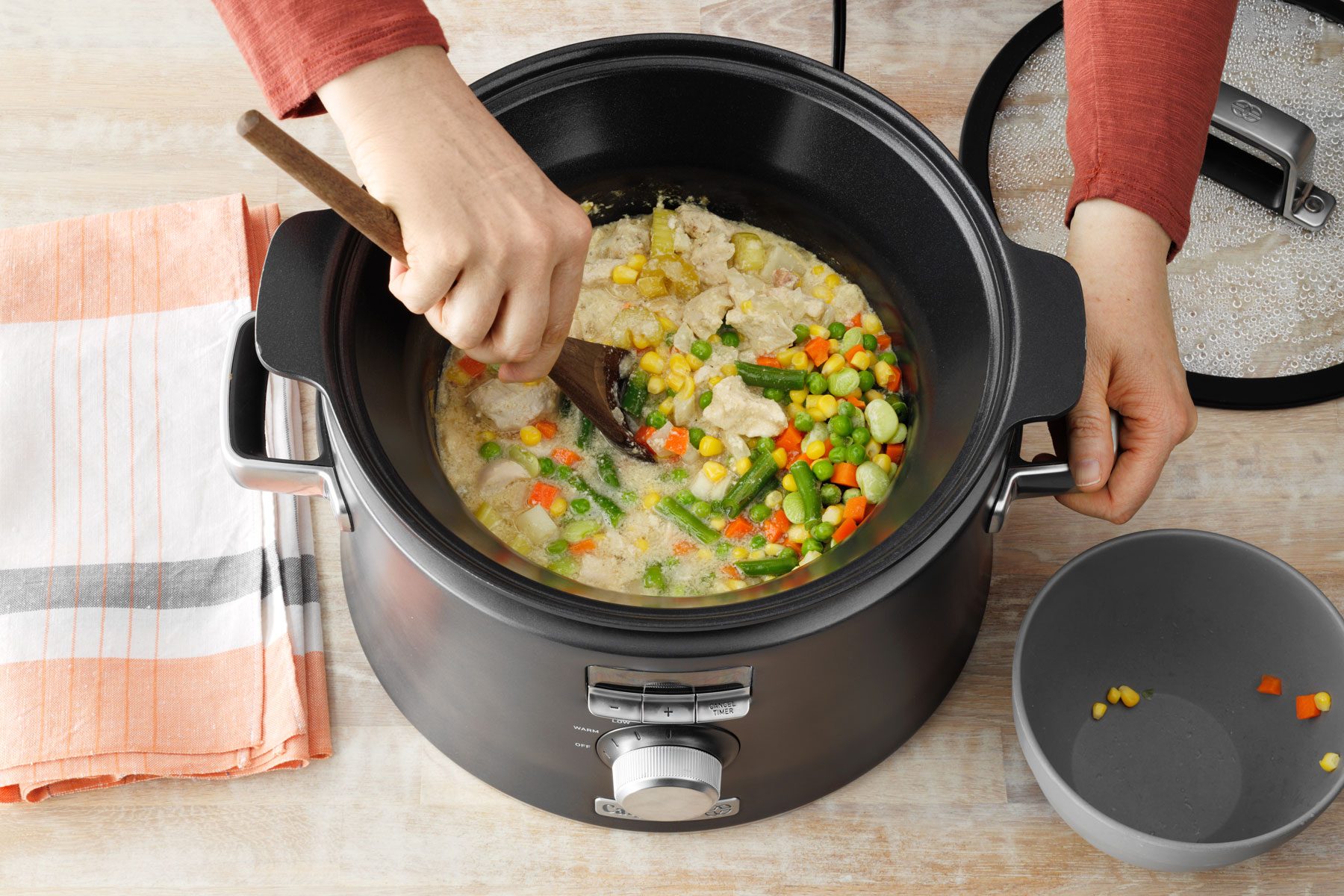 A person stirs a mix of chicken, vegetables, and broth in a slow cooker with a wooden spoon. A folded striped cloth napkin and an empty gray bowl are placed next to the slow cooker on a wooden surface. The lid of the slow cooker is partially visible.