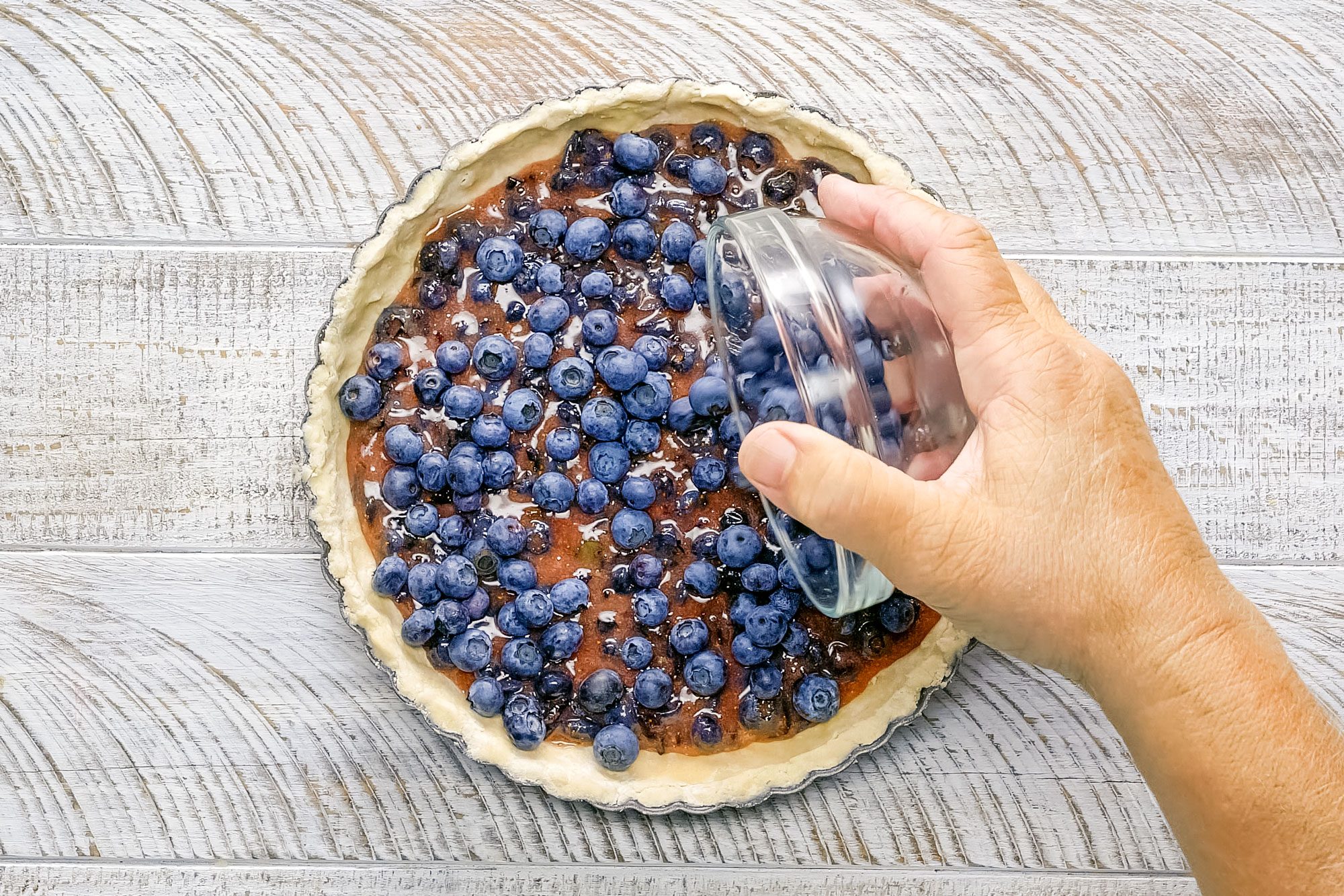 Pouring Blueberry Tart Filling on pressed dough in tart pan on wooden surface