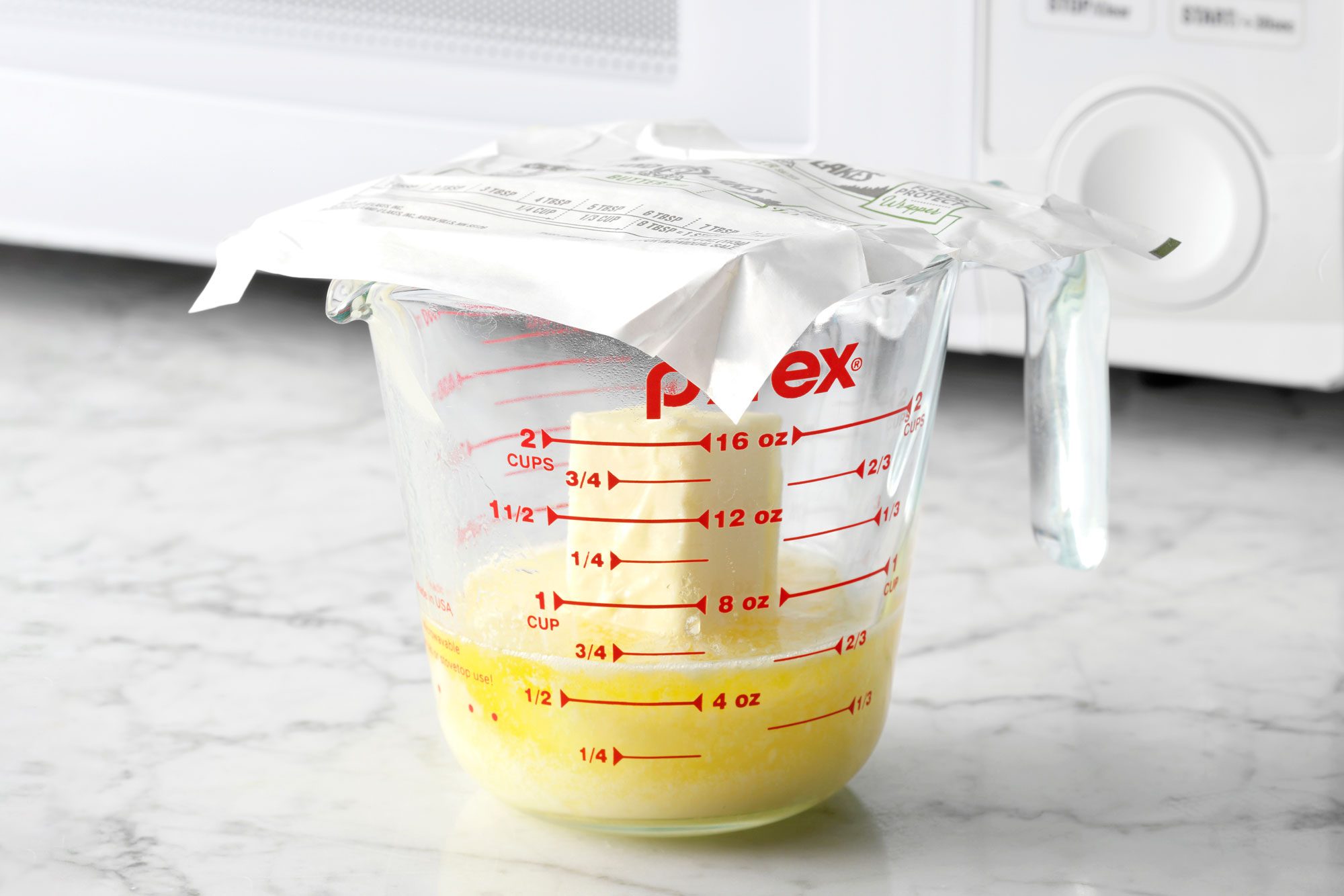 Melting Butter in a glass measuring cup With a butter Wrapper on top sitting on the counter, a microwave is nearby in the background