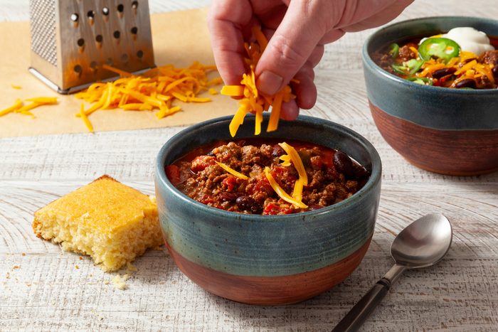 A person putting cheese into a bowl of chili con carne