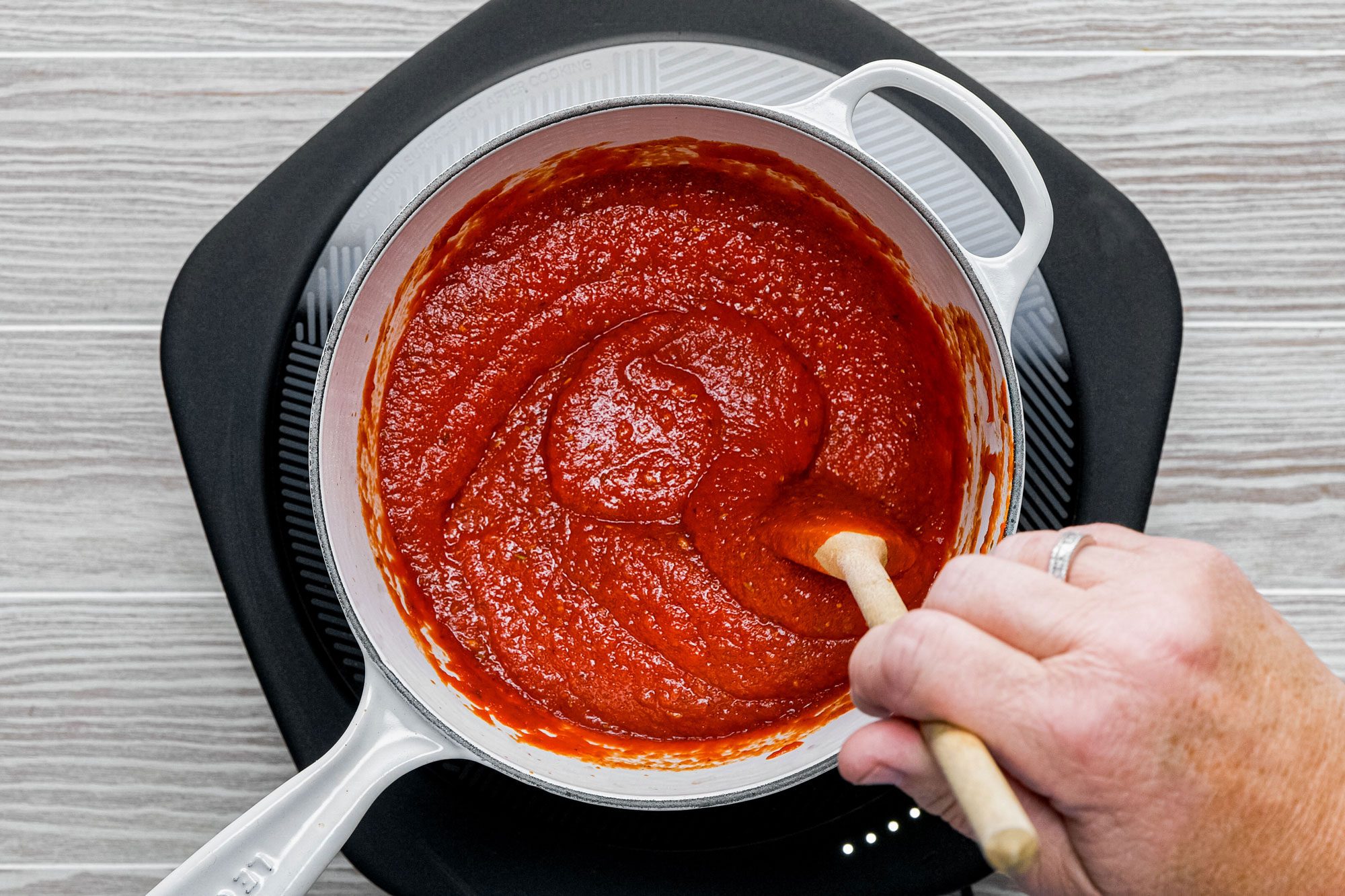 A hand stirs pizza sauce with a spatula in a saucepan on an induction cooktop on wooden surface