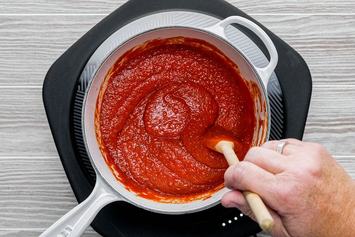 A hand stirs pizza sauce with a spatula in a saucepan on an induction cooktop on wooden surface