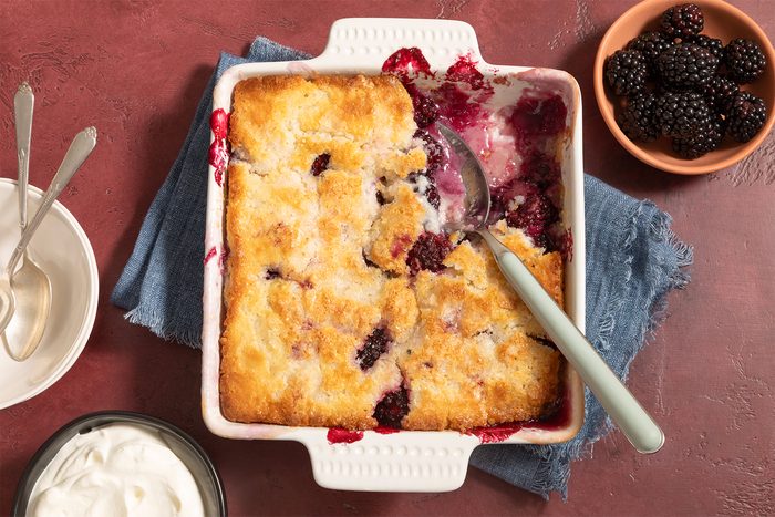 A partially served berry cobbler in a square white baking dish, with a silver serving spoon resting inside. Beside the dish are bowls containing fresh blackberries and whipped cream, along with two spoons resting on a light-colored plate. A blue cloth is underneath.