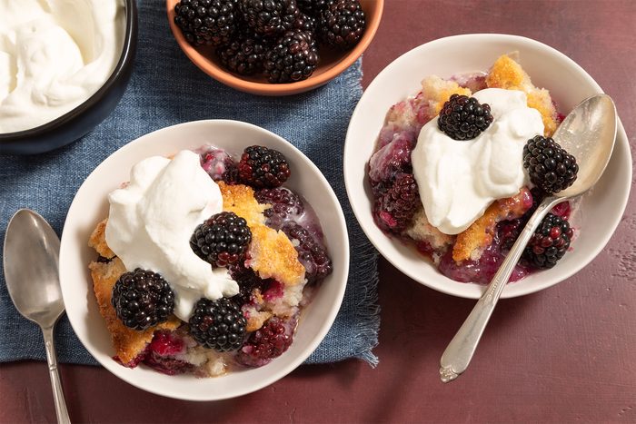 Two bowls of blackberry cobbler topped with whipped cream, accompanied by spoons, are displayed on a blue cloth. A bowl of fresh blackberries and a bowl of whipped cream are also present nearby on a red surface.