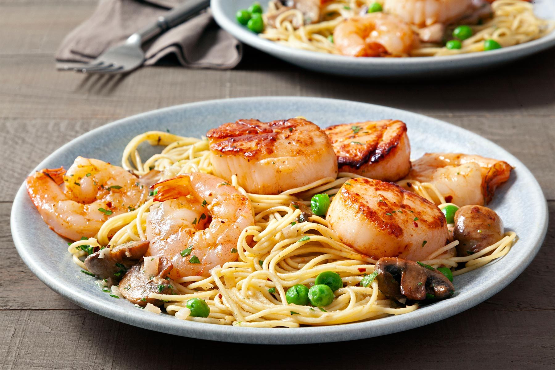 Table view shot of Sauteed Scallops & Shrimp Pasta; served in two plates; sprinkle with parsley; fork; napkin; wooden background;