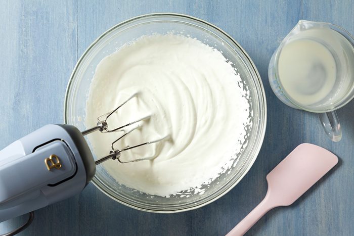 beating cream In a large glass bowl using a hand mixer on a textured light blue surface